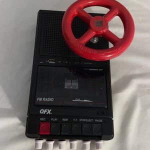 May include: A black FM radio with a red steering wheel-shaped knob. The radio has buttons for record, play, rewind, fast forward, stop/eject, and pause. The text "OFX" is visible on the radio.