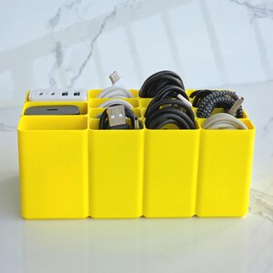 May include: A vibrant yellow cable tidy with several compartments. It contains various cables, a power strip, and a small electronic device. Cables are black, white, and grey. The organiser is designed to keep cords neat and tidy.