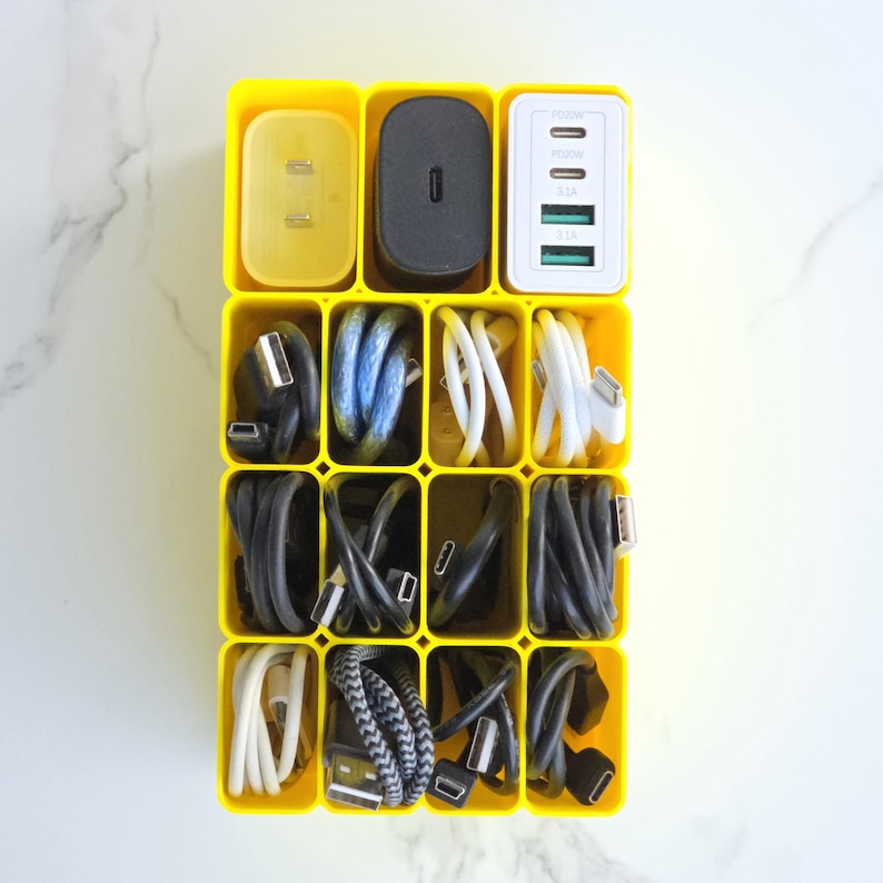 May include: A yellow cable organiser containing a variety of chargers and cables. The top row features a yellow plug, a black charging block, and a white charger with multiple ports. The lower compartments are filled with coiled black, blue, and white cables.