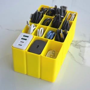 May include: A bright yellow cable organizer with multiple compartments, filled with various black, white, and blue charging cables and power adapters. The organizer includes a white multi-port USB charger and a black power adapter.