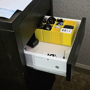 May include: An open drawer with a yellow organiser holding black, white, and blue charging cables and a multi-port USB charger. The drawer is part of a dark brown desk or dresser. Other items are visible in the drawer.