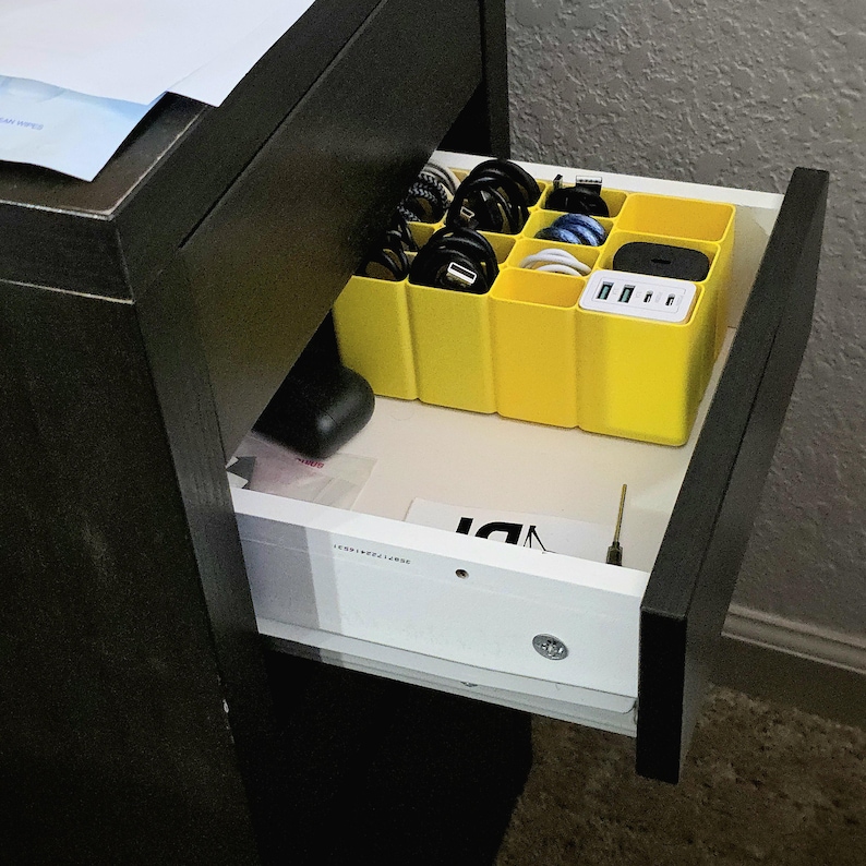 May include: An open drawer with a yellow organiser holding black, white, and blue charging cables and a multi-port USB charger. The drawer is part of a dark brown desk or dresser. Other items are visible in the drawer.