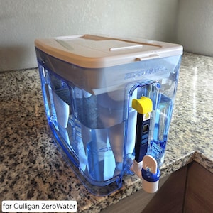 May include: A clear, blue ZeroWater water filter pitcher with a white lid and a yellow and black TDS meter. The pitcher is filled with water and sits on a granite countertop. The text "ZeroWater" is visible on the top.