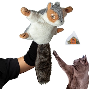 May include: A soft squirrel toy in shades of grey, white, and brown is held aloft. The toy has a fluffy tail and outstretched arms. A small bag of catnip infusion is visible. A grey cat reaches towards the toy.