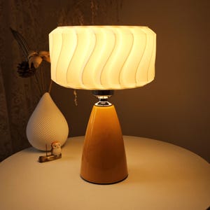 Könnte beinhalten: Eine moderne Tischlampe mit gelbem Sockel und weißem, wellenförmigem Schirm. Die Lampe ist eingeschaltet und strahlt ein warmes, einladendes Licht aus. Das Design ist modern und eignet sich für ein Wohn- oder Schlafzimmer. Die Lampe steht auf einem weißen Tisch.