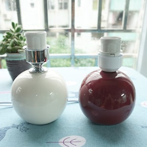 May include: Two ceramic table lamps with round white and burgundy shades. The lamps have a silver metal base and a white plastic socket.