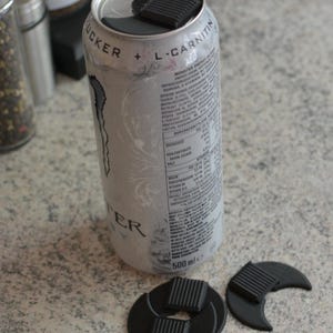 Könnte beinhalten: Eine silberne Monster Energy-Dose mit schwarzen Kunststoff-Dosenaufsätzen. Die Dose hat weiße und graue Grafiken und Text. Die Dosenaufsätze sind schwarz und haben ein geriffeltes Design. Die Dose ist 500 ml.
