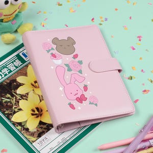 May include: A pink planner with a cartoon bunny and floral design. The planner has a magnetic closure and is surrounded by pens, a plush frog, and a Japanese language notebook.