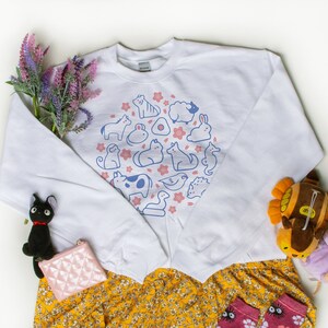 May include: White sweatshirt with a blue and pink graphic of various animals and flowers. The sweatshirt is paired with a yellow floral skirt and pink socks with black eyes and flowers.