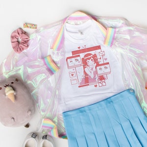 May include: A white t-shirt with a pink anime girl graphic, a light blue pleated skirt, a white iridescent jacket with rainbow trim, a pink hair scrunchie, a brown cat plush toy, and a Japanese manga book.