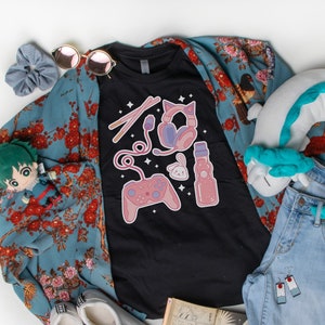 K&ouml;nnte beinhalten: Ein schwarzes T-Shirt mit einem Grafikdesign einer Videospielkonsole, Kopfh&ouml;rern, einem Kaninchen und einer Flasche Limonade. Das Shirt liegt auf einem blauen, floralen Kimono, einer blauen Jeans, einem wei&szlig;en Kissen mit blau-wei&szlig;em Design und einem Manga-Buch.