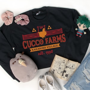 May include: Black sweatshirt with a red and yellow logo that says "Cucco Farms Kakariko Village est. 1998". The logo features a chicken in a fenced-in area with a triangle design. The sweatshirt is laid out on a white surface with a pair of white sneakers, a pair of blue jeans, a small green cartoon character, and a small grey cat plush toy.