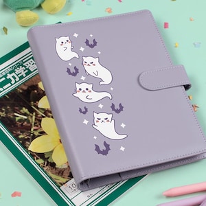 May include: A lavender-colored planner with a cat and bat design. The planner has a snap closure and is lying on a light green surface with a textbook and pens. A plush frog toy is in the upper left corner.