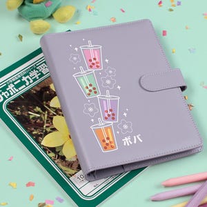 May include: A light purple binder with a snap closure, decorated with a graphic of colorful boba tea cups and Japanese text. A plush frog toy and a Japanese textbook are also in the image.