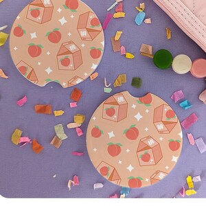 May include: Two peach-themed car coasters with a light peach background, featuring illustrations of peaches, milk cartons, and stars. The coasters are on a purple surface with colorful confetti and a pink purse.