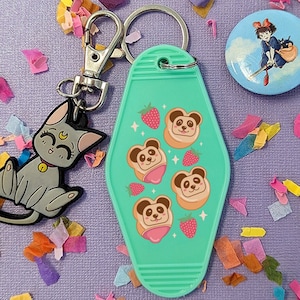 May include: A teal blue motel key tag keychain with a design of panda bears with strawberry and pink frosting on top. The keychain is attached to a silver split ring with a black cat charm.