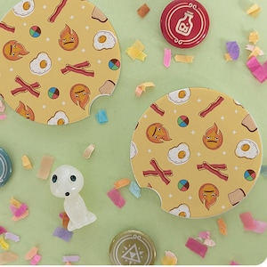 May include: Two yellow car coasters with a breakfast food pattern, including bacon, eggs, and toast, and various emoji faces. Also, several decorative pins and a small figurine are scattered on a light green surface.