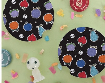 May include: Two black and white round coasters with a repeating pattern of colorful cartoon fruits and skulls. The coasters are on a green surface with confetti.