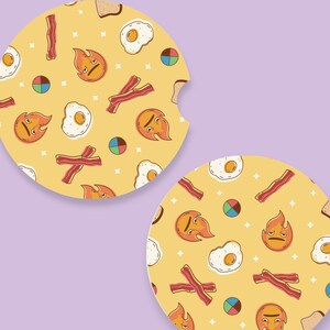 May include: Two round car coasters with a yellow background and a repeating pattern of fried eggs, bacon strips, toast, and flame-shaped faces. The coasters have a small notch for easy removal.