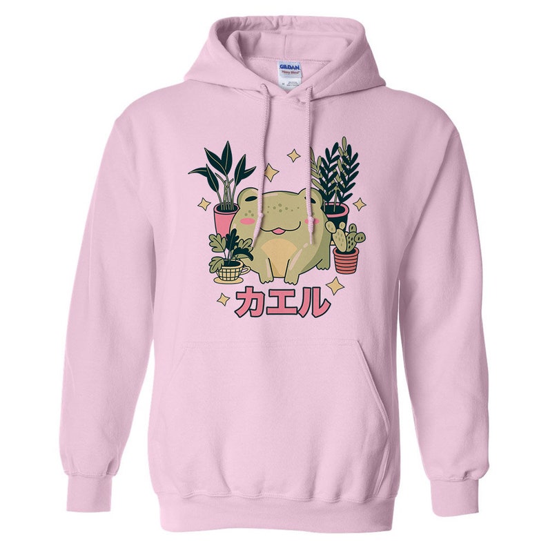 May include: A pink hoodie with a green frog sitting among potted plants. The frog is smiling and the text "カエル" is printed below the frog.