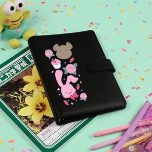 Peut inclure: Agenda noir avec un design fantaisiste repr&eacute;sentant un lapin rose, un ours brun, des cupcakes, des roses et des fraises. L'agenda a une fermeture magn&eacute;tique et est entour&eacute; d'autres articles de papeterie.