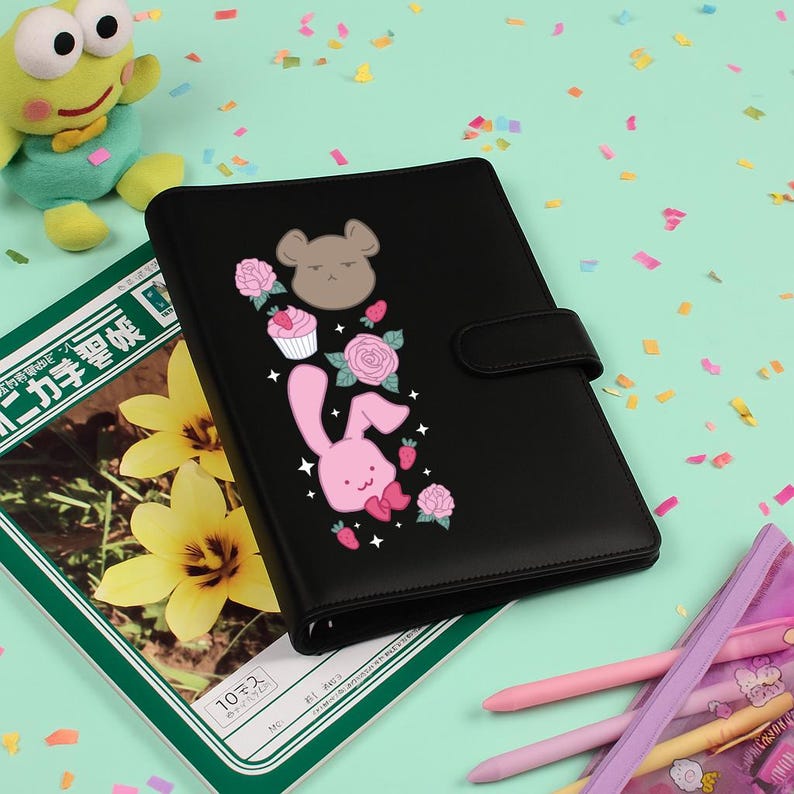 Peut inclure: Agenda noir avec un design fantaisiste repr&eacute;sentant un lapin rose, un ours brun, des cupcakes, des roses et des fraises. L'agenda a une fermeture magn&eacute;tique et est entour&eacute; d'autres articles de papeterie.