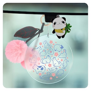 May include: A clear round charm with a white outline of various animals and pink flowers. The charm is attached to a gold chain with a black and white panda bear keychain.
