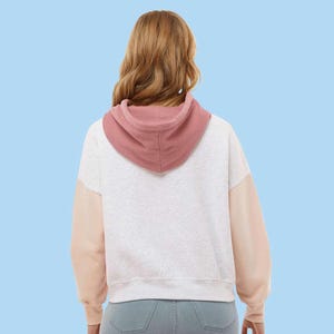 May include: A white and gray hoodie with a pink hood and peach-colored sleeves. The back view of the hoodie is shown against a light blue background. The hoodie is made of a soft material.