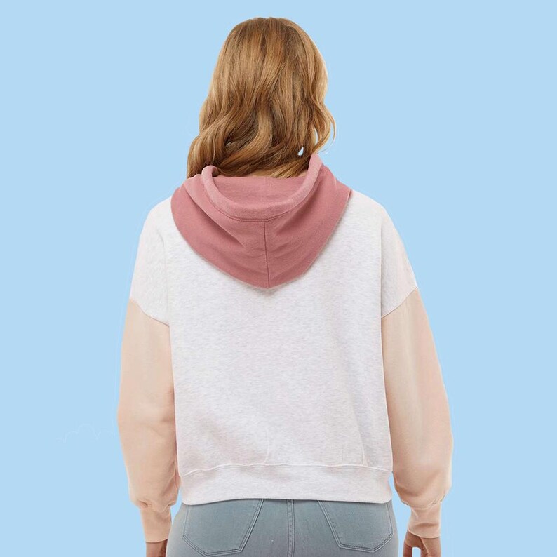 May include: A white and gray hoodie with a pink hood and peach-colored sleeves. The back view of the hoodie is shown against a light blue background. The hoodie is made of a soft material.