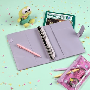 Peut inclure: Un agenda lilas avec un classeur argent&eacute;, ouvert pour r&eacute;v&eacute;ler des poches et un stylo rose. Un livre vert avec du texte japonais, une peluche grenouille et une trousse transparente avec des articles de papeterie sont &eacute;galement pr&eacute;sents. Le fond est vert clair.