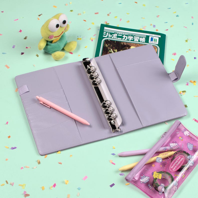 Peut inclure: Un agenda lilas avec un classeur argent&eacute;, ouvert pour r&eacute;v&eacute;ler des poches et un stylo rose. Un livre vert avec du texte japonais, une peluche grenouille et une trousse transparente avec des articles de papeterie sont &eacute;galement pr&eacute;sents. Le fond est vert clair.