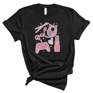 K&ouml;nnte beinhalten: Schwarzes T-Shirt mit einer rosa und blauen Illustration eines Videospiel-Controllers, Kopfh&ouml;rern, einer Flasche Limonade und einem kleinen Kaninchen. Die Illustration ist von kleinen wei&szlig;en Sternen umgeben.