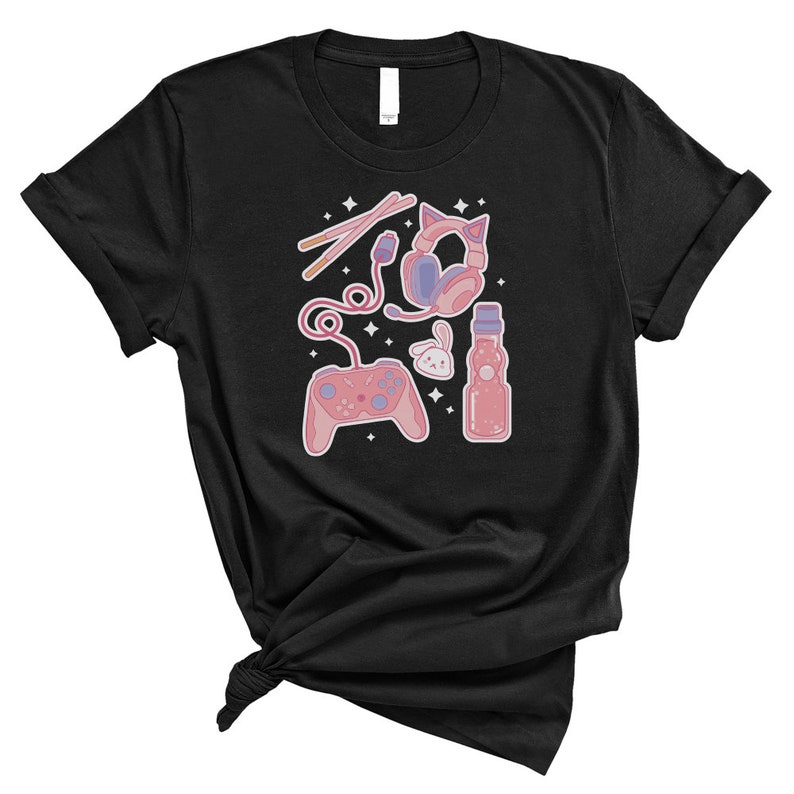 K&ouml;nnte beinhalten: Schwarzes T-Shirt mit einer rosa und blauen Illustration eines Videospiel-Controllers, Kopfh&ouml;rern, einer Flasche Limonade und einem kleinen Kaninchen. Die Illustration ist von kleinen wei&szlig;en Sternen umgeben.