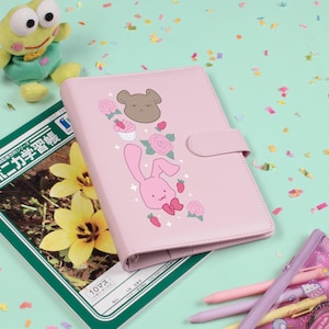 May include: A pink planner with a cartoon bunny and floral design. The planner has a magnetic closure and is surrounded by pens, a plush frog, and a Japanese language notebook.