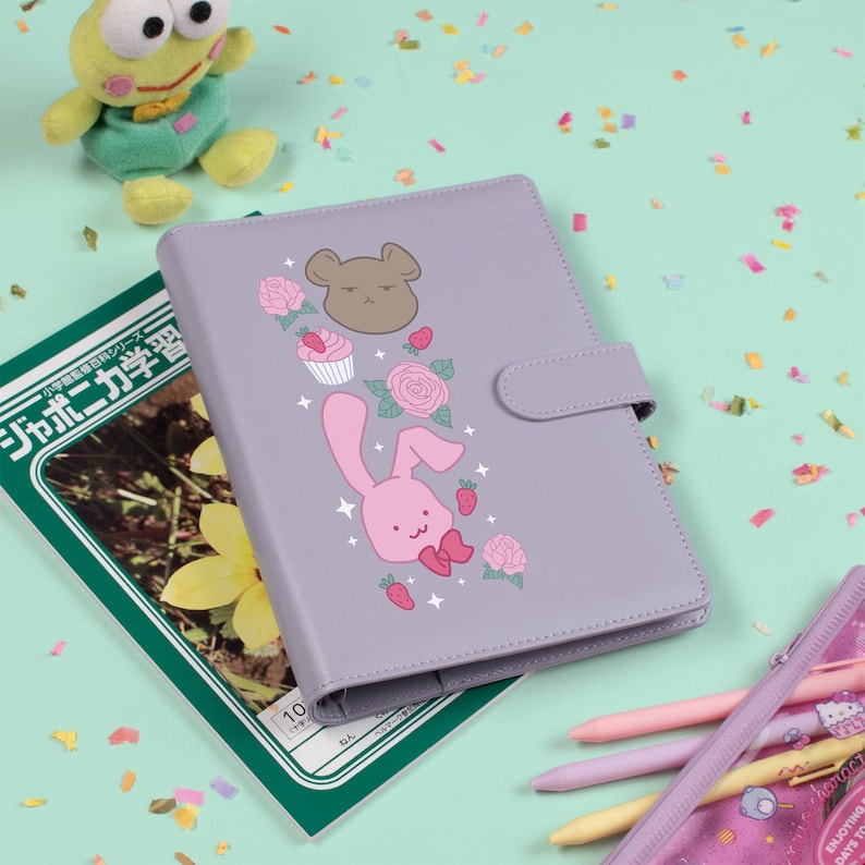 Peut inclure: Un agenda violet clair avec un dessin de lapin, d'ours, de cupcake et de rose. L'agenda a une fermeture &agrave; bouton-pression et se trouve &agrave; c&ocirc;t&eacute; d'un manuel japonais et d'une trousse avec des stylos color&eacute;s.