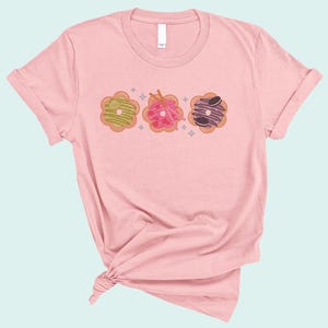 May include: A pink t-shirt with a graphic of three donuts with different toppings. The donuts are in a row and have a light pink, green, and purple frosting.