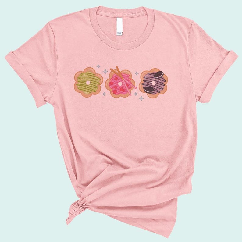 May include: A pink t-shirt with a graphic of three donuts with different toppings. The donuts are in a row and have a light pink, green, and purple frosting.