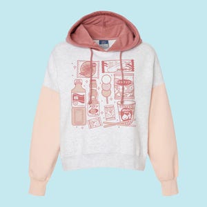 May include: A light gray hoodie with a pink hood and sleeves. The front of the hoodie features a graphic of various Japanese snacks and drinks in a pink color. The hoodie has a drawstring and a tag on the hood.