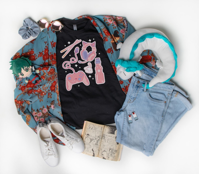 K&ouml;nnte beinhalten: Ein schwarzes T-Shirt mit einem Grafikdesign einer Videospielkonsole, Kopfh&ouml;rern, einem Kaninchen und einer Flasche Limonade. Das Shirt liegt auf einem blauen, floralen Kimono, einer blauen Jeans, einem wei&szlig;en Kissen mit blau-wei&szlig;em Design und einem Manga-Buch.