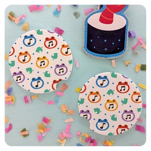 May include: Two white round drink coasters with a colorful frog and musical note pattern. The coasters are on a light blue background with confetti.