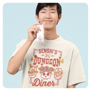May include: A person wearing a beige t-shirt with a graphic design that says "Senshi's Dungeon Diner". The graphic is in red and yellow with a skull and crossbones in the center. The person is holding a small carton of milk.