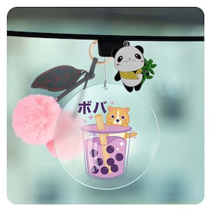 May include: A round, clear, acrylic car charm with a cartoon image of a dog drinking boba tea. The text "ボバ" is written in Japanese on the charm. A pink fuzzy pom-pom and a panda bear charm are attached to the charm.