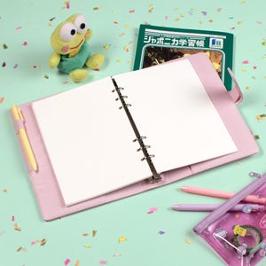 Peut inclure: Un classeur rose clair avec un cahier pointill&eacute; ouvert, un stylo jaune et un manuel japonais vert. Une peluche grenouille et une trousse transparente avec des stylos roses et violets sont &eacute;galement visibles. Le fond est vert clair avec des confettis.