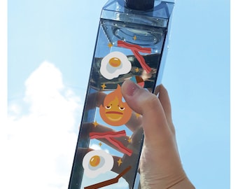 May include: A clear plastic milk carton-shaped water bottle with a black lid. The bottle has various stickers on it, including a cartoon fire character, bacon, and eggs.