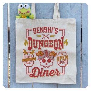 May include: A white canvas tote bag with a red and yellow graphic design that says "Senshi's Dungeon Diner". The graphic includes a skull with horns and a crown, a sword and fork, and the words "Fresh Monsters Daily!" and "Open 24/7!"