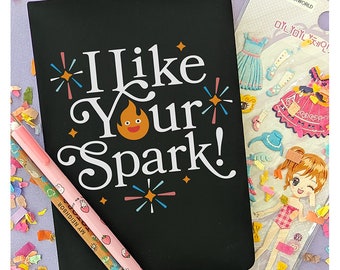 May include: A black notebook with the text "I Like Your Spark!" in white lettering. The notebook has a small orange flame with a smiling face on it. The notebook is surrounded by confetti and two pencils.
