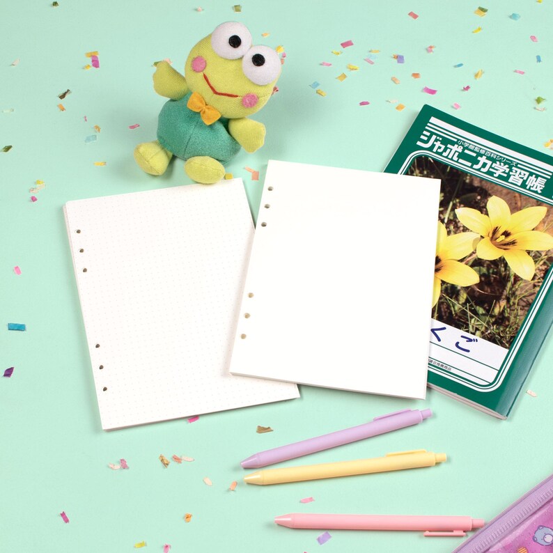 Peut inclure: Un ensemble de papeterie comprenant une peluche grenouille, du papier &agrave; grille de points vierge, un livre d'apprentissage de la langue japonaise et trois stylos pastel. Les articles sont dispos&eacute;s sur une surface vert clair avec des confettis color&eacute;s.