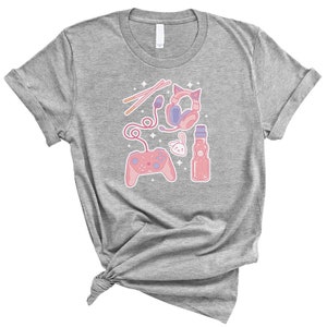 K&ouml;nnte beinhalten: Ein hellgraues T-Shirt mit einem pinkfarbenen und violetten Design, das einen Videospielcontroller, Kopfh&ouml;rer, eine Flasche Ramune und Essst&auml;bchen zeigt.