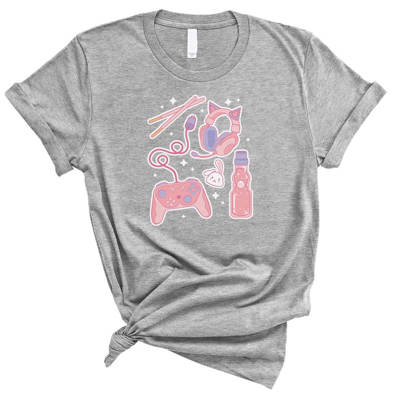 K&ouml;nnte beinhalten: Ein hellgraues T-Shirt mit einem pinkfarbenen und violetten Design, das einen Videospielcontroller, Kopfh&ouml;rer, eine Flasche Ramune und Essst&auml;bchen zeigt.