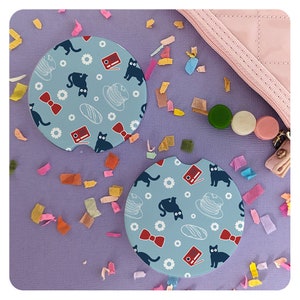 May include: Two blue and white ceramic coasters with a black cat, a red bow, a radio, a hat, and a baguette design. The coasters are on a purple surface with colorful confetti.